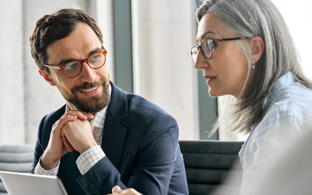 Two people that appear to be talking to others in the room. The woman in the foreground is speaking and the man in the background is looking at her.