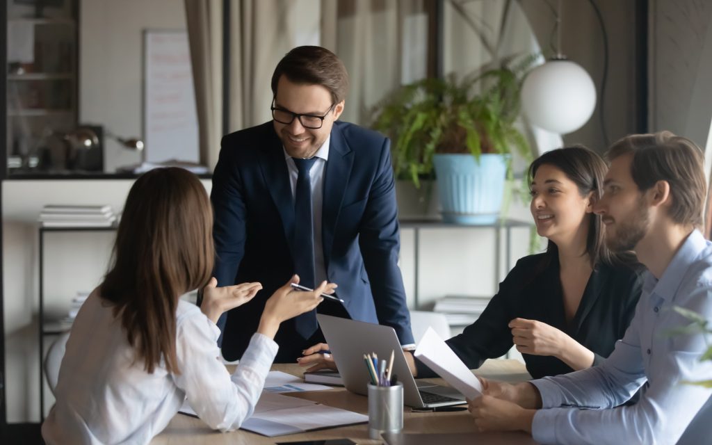 A group of people having a conversation around a table with laptops, pencils, and note paper.