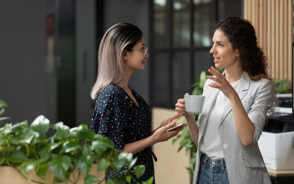 Two people having a conversation in an office. One person has their phone in their hand and the other has a cup of coffee.